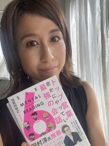 Close-up of a smiling woman holding a pink-and-white Japanese magazine partially covering her chest, indoors by sheer curtains.