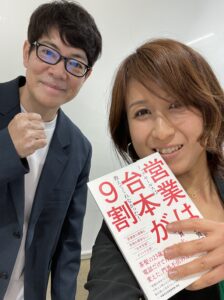 Two people posing for a selfie: a man in glasses and a blazer on the left, and a smiling woman holding a white book with red Japanese text on the right.