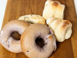 Two light brown bagels with holes sit on a wooden board, with several small dinner rolls in the background.